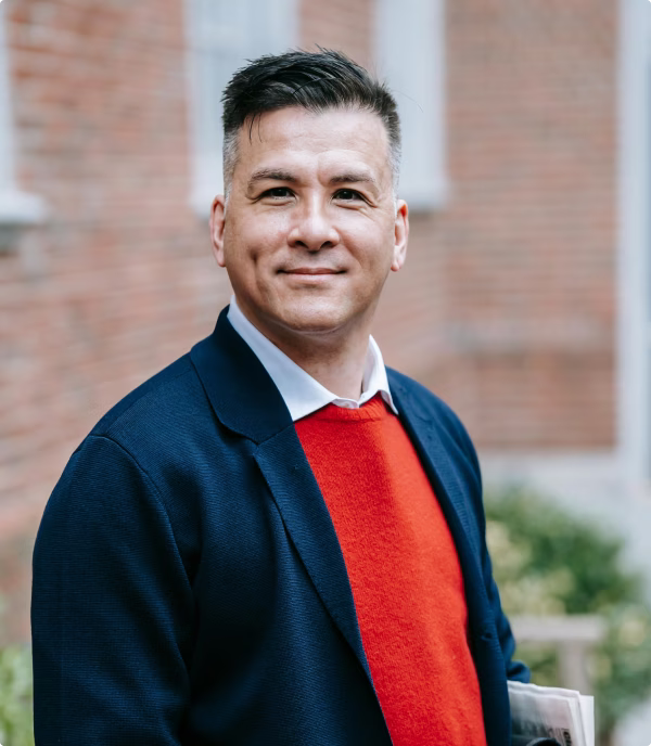 Man with short dark hair wearing a navy blazer and red sweater standing outdoors against a blurred brick wall background.